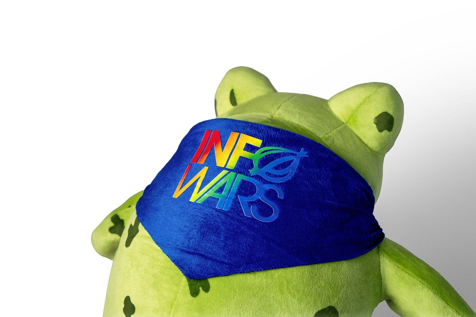 A plush InfoWars Gay Frog Toy with a bright green body, yellow belly, a blue handkercheif bow tied around its neck, and colorful rainbow-print hangtag on a white background.