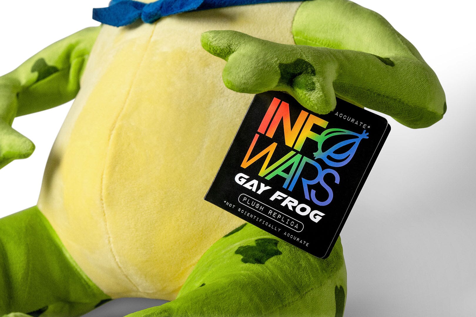 Close-up of a plush InfoWars Gay Frog collectible with green limbs and torso, a pale yellow belly, and a blue handkercheif tied around his neck. A black hangtag reading 'Infected Wars Gay Frog' is attached near the hand.
